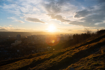 Sunset over a cityscape viewed from a hillside, with buildings and mountains visible in the distance under a partly cloudy sky