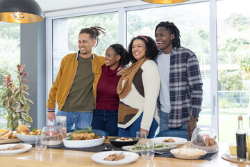 Diverse friends linking arms, standing at dining table at home with roasted chicken platter