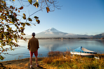 A man stands on the grassy shore of a serene lake Kawaguchi, gazing toward the snow-capped peak of Mount Fuji as a small boat rests nearby under a clear blue sky. © Agnieszka Gaul