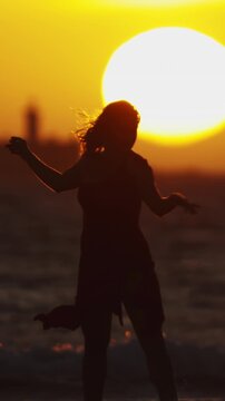 Dark silhouette of dancing young woman on the seashore during bright orange sunset