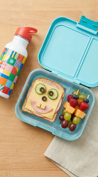 An organized overhead view of a child&rsquo;s bento lunchbox with a funny face sandwich, fruit skewers, and a colorful water bottle on a clean kitchen counter.
