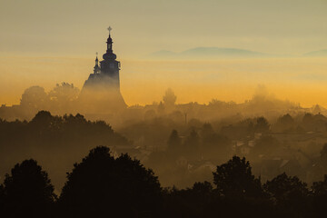 Wiejski kościół z wysoką wieżą we mgle w górskim krajobrazie w blasku wschodzącego słońca © piotr