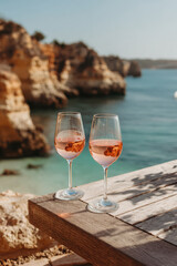 Fototapeta premium Two chilled glasses of rosé wine on a rustic wooden table overlooking a rocky Mediterranean bay with turquoise water in warm golden summer sunlight