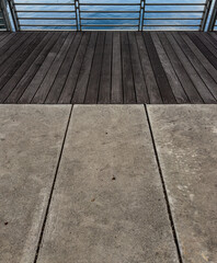 Fototapeta premium A concrete walkway meets a dark wooden deck with visible screws and a metal railing overlooking calm blue water creating a structured peaceful waterfront scene