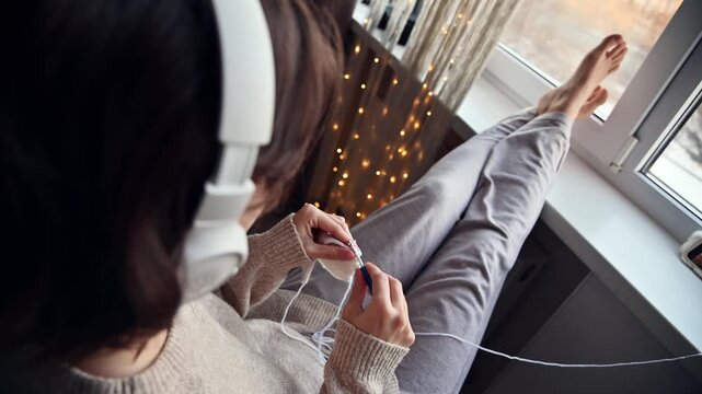 Young woman with headphones sits on a windowsill, engaged in a creative activity with yarn and a crochet hook, warm lighting and decorative lights in the background
