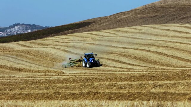 blue tractor pulling cultivator kicking up dust golden wheat field distant village rolling hills agriculture farming concept rural landscape machinery dry summer nature food production scenic