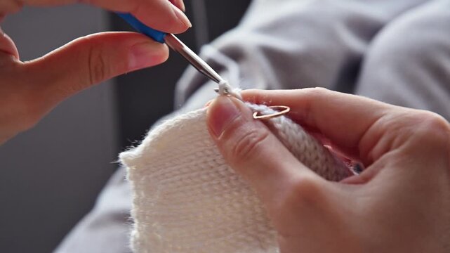 Hands of a woman using a crochet hook to attach a stitch to a white knitted fabric, showcasing a detailed crafting process in a cozy indoor setting