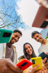Obraz premium Vertical shot of young group of people using phones devices standing together in circle outdoors. Millennial friends addicted to social media app, betting or playing video game on platform online.
