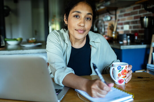 Woman working from home at kitchen table with laptop and coffee
