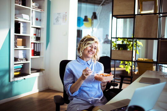 Smiling woman eating lunch at office desk with laptop