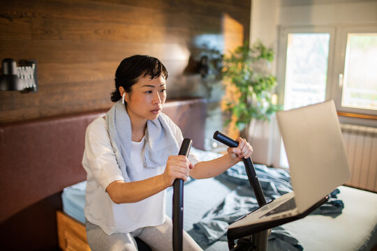 Focused woman on exercise bike with laptop in bedroom