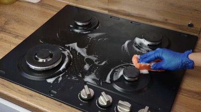 A man cleaning a gas stove, gloved hands with a sponge and foam, close-up. Concept of house cleaning, household chores