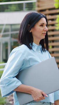 Confident Caucasian woman in sunglasses and casual clothes is looking at the camera while holding a laptop outdoors, standing in front of glass modern building in park setting. Slow otion