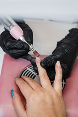 Fototapeta premium A professional nail technician in black gloves uses an electric nail drill to perform a hardware manicure on a client's fingernails over a dust collector in a studio.