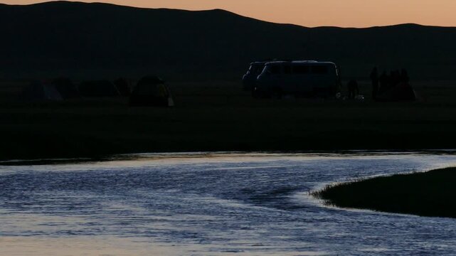 A night river against the background of low mountains and a tourist camp under a pink sunset sky. Summer warm night.
