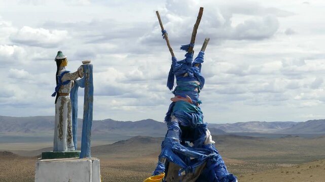Santmargats, MONGOLIA - July 18, 2017. Tied with blue ritual scarves, the wooden top of oboo  (ovoo, ritual heap of stones) and a woman sculpture with a scarf in her hands against the cloudy sky.