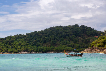 Obraz premium Coastal scene in Thailand, near Krabi. A traditional wooden long-tail boat is floating on the vibrant turquoise Andaman Sea. A sloping hill is completely covered in lush green tropical vegetation.