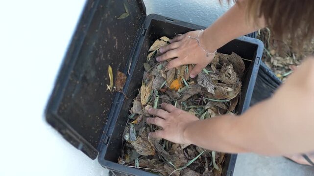 Close-up of a woman's hands stirring leaves inside a black urban compost bin made from recycled plastic.