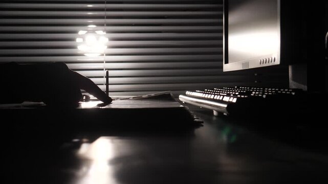 Hand silhouette points at a document on a dim office desk beside keyboard and monitor, moody low key scene evoking investigation, secrecy, data review, and late night work