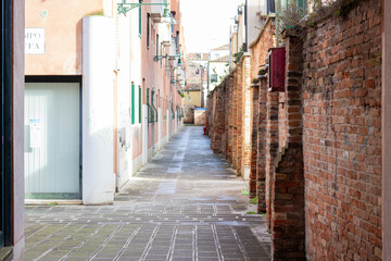 Obraz premium Narrow cobblestone alleyway lined with brick walls and buildings, featuring green window shutters and a clear blue sky in a historic European city