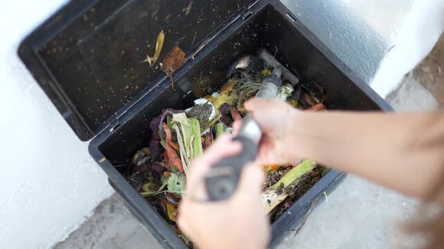 Close-up of a woman's hands mixing organic waste and leaves with a rake inside a black recycled plastic urban compost bin.