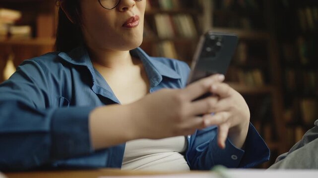 Happy Asian female student procrastinating in library, scrolling cellphone