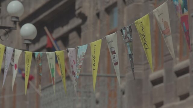 A string of decorative, colorful, triangular pennant flags with various unique illustrations gently flutters in the breeze against a blurred background of a city building and street lamps.

