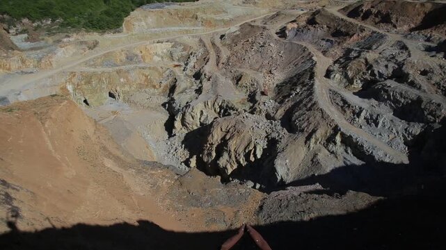 Aerial view looking into a deep open pit mine. A slow camera tilt reveals the rugged, excavated landscape with winding dirt tracks, exposed rock layers, and steep drop-offs.

