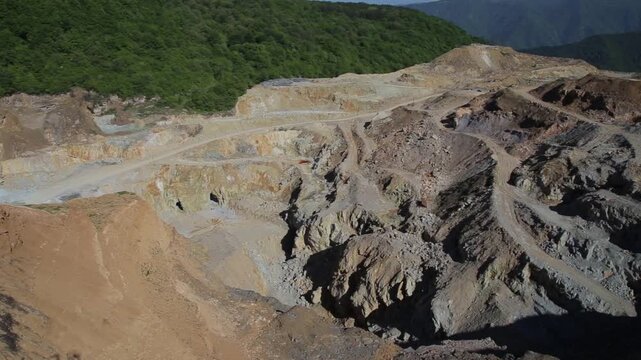 Aerial view looking into a deep open pit mine. A slow camera tilt reveals the rugged, excavated landscape with winding dirt tracks, exposed rock layers, and steep drop-offs.

