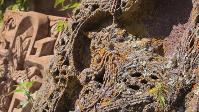 Close up view of an ancient Armenian khachkar cross stone. The porous textured rock surface features intricate carvings and is covered with orange lichen and tiny green plants.

