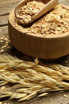 Oat flakes in bowl and panicles on wooden table, closeup
