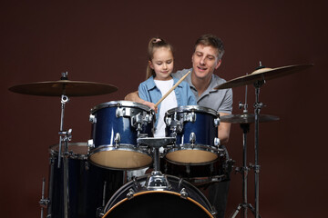 Little girl learning how to play drums with music teacher on brown background