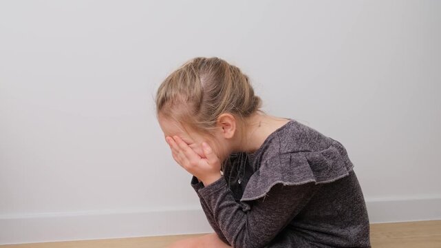 Little Girl Cries and then Calmly Looks Up During a Reflective Emotional Moment, Sitting on a Wooden Floor by a White Wall