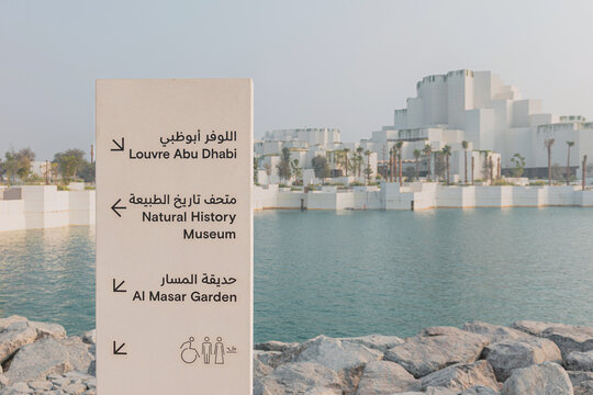 6 December 2025, Abu Dhabi, UAE: Directional signage indicating Louvre, Natural History Museum, and Al Masar Garden, with the modern architecture and tranquil water