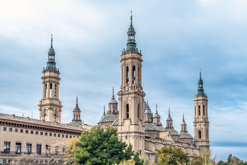 Obraz premium Beautiful side view of the Basilica del Pilar with multiple towers and colorful tiled domes in Zaragoza, Spain. This landmark showcases magnificent baroque architecture along with green trees