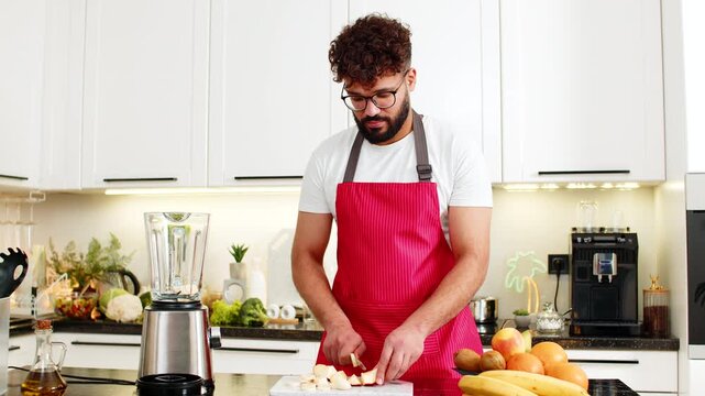 Arabian man drops apple and banana pieces into blender in kitchen preparing fresh smoothie for healthy nutrition at home. Young guy dances to music while making fitness breakfast drink with fruit mix.