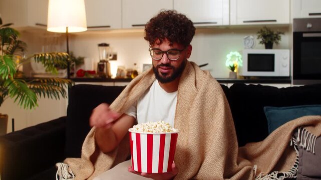 Arabian man at home watches movie on tv holds popcorn laughs comments enjoys calm evening. Egyptian guy on sofa streams show in blanket talks to screen smiles reacts to funny moments at night indoors.