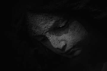 Fototapeta premium Dark stone statue face emerging from shadows, symbol of sorrow, silence, grief, and spiritual contemplation.