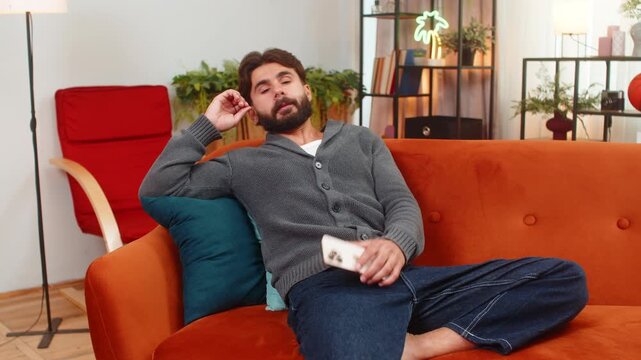 Indian man at home naps near smartphone sudden notification sound wakes up startled reads message important news. Arabian guy on sofa looks at screen quick breath reacts with surprise alert very fast