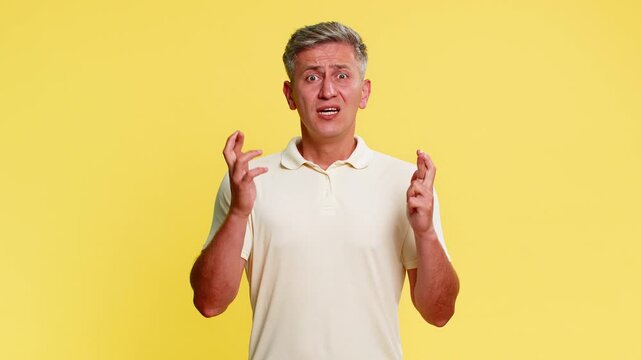 Middle-aged man crossing fingers with hopeful face expecting positive result, reacting with clear disappointment visible frustration. Young guy showing failure emotion isolated on yellow background.