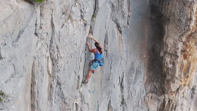 AERIAL: Focused woman in blue outfit is captured in moment of intense concentration while climbing a technical section of a limestone rock face. She uses chalk to improve friction and better grip.