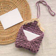 Purple handmade crochet book sleeve with blank white card for copy space on wicker tray and linen background.