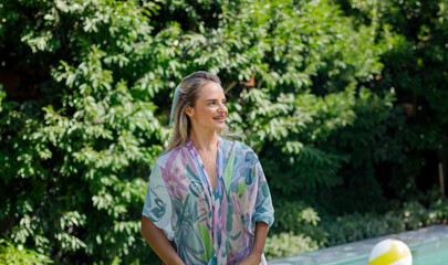 Woman standing poolside in sunlit garden wearing pastel floral cover-up near inflatable beach ball