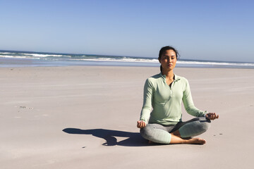 Asian woman meditating cross-legged on sand by ocean, in pullover leggings, copy space