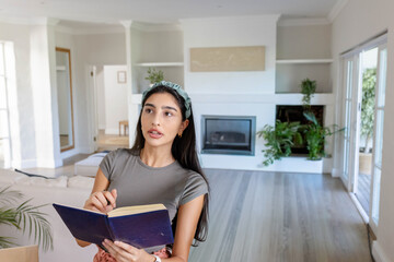 Fototapeta premium Indian woman standing and holding navy-blue notebook and pen, looking upward in living room