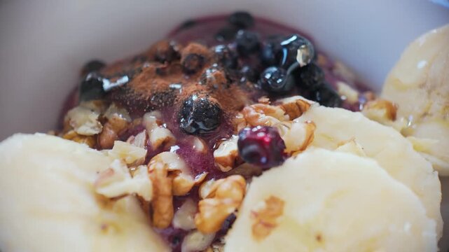 Macro slow motion cinnamon sprinkle over oatmeal with fruit and nuts, aromatic healthy breakfast finishing detail