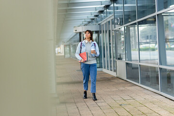 Mid-adult woman walking along walkway beside glass facade holding red folder, coffee cup, backpack