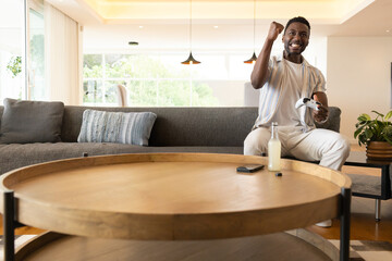 African American man sitting right of sofa holding white gamepad by wooden coffee table, copy space © wavebreak3