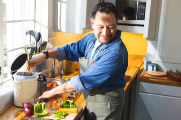 Mature Asian man standing in sunlit kitchen wearing denim shirt green apron, cutting bell pepper © wavebreak3