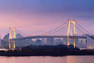 Obraz premium Tokyo, Japan – Evening cityscape featuring Rainbow Bridge illuminated over Tokyo Bay with Tokyo Tower visible in the background, showcasing modern skyline and urban architecture at dusk in Tokyo, 